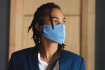 Retrato de hombre de raza mixta con máscara facial de pie en el vestíbulo del hotel con bolso de hombro. viaje de negocios hotel durante coronavirus covid 19 pandemia. - foto de stock