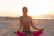Mujer caucásica en la playa practicando yoga sentada en meditación. salud y bienestar, relajarse en la playa al amanecer. - foto de stock