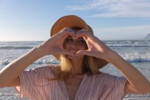 Кавказька жінка у капелюсі робить серцеву форму, дивлячись на камеру і посміхаючись на пляжі. Здоровий вільний час на відкритому повітрі біля моря. — стокове фото
