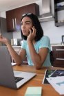 Змішана раса трансгендерна жінка працює вдома, використовуючи ноутбук, що розмовляє на смартфоні. перебування вдома в ізоляції під час карантину . — стокове фото