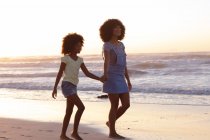 Посміхаючись афроамериканською матір'ю і дочкою, які ходять і тримаються за руки на пляжі. Здоровий вільний час на відкритому повітрі біля моря. — стокове фото