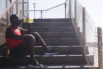African american man exercising, resting, drinking water on stairs on sunny day. healthy outdoor lifestyle fitness training. - foto de stock