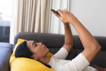 Glückliche Transgender-Frau mit gemischter Rasse entspannt sich im Wohnzimmer auf der Couch liegend und macht Selfies. Isolationshaft während der Quarantäne. — Stockfoto