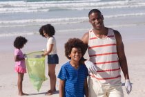 African american parents with two children collecting rubbish from the beach smiling. family eco beach conservation. — Photo de stock