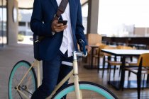 Seção média do macho sentado na bicicleta fora do café usando smartphone. nômade digital, para fora e sobre na cidade. — Fotografia de Stock
