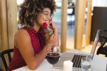 Mujer de raza mixta sonriendo sentada en la mesa en el restaurante teniendo videollamada. amigos hablando entre sí en línea sentado en un restaurante. - foto de stock