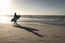 Mulher sênior andando na praia segurando prancha de surf olhando para o mar. saúde e bem-estar, reforma activa. — Fotografia de Stock