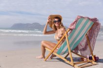 Усміхнена бікіні жінка сидить на стільці, дивлячись на камеру на пляжі. здоровий відпочинок на відкритому повітрі біля моря . — стокове фото
