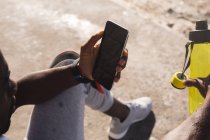 Afrikanisch-amerikanischer Mann, der an sonnigen Tagen am Strand Sport treibt, sich ausruht und sein Smartphone benutzt. gesundes Outdoor-Fitness-Training. — Stockfoto