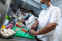 Diverse männliche und weibliche Profiköche bereiten Gemüse mit Gesichtsmasken zu. Arbeit in einer belebten Restaurantküche während der Coronavirus-Pandemie 19. — Stockfoto