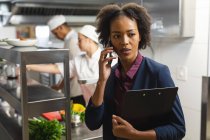 Mixed Race Kitchen Manager telefoniert mit professionellen Köchen im Hintergrund. Arbeit in einer belebten Restaurantküche. — Stockfoto