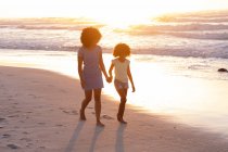 African american mother and daughter walking and holding hands at the beach. healthy outdoor leisure time by the sea. - foto de stock
