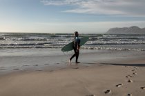 Mujer afroamericana mayor llevando tabla de surf mientras camina por la playa. viaje vacaciones retiro estilo de vida concepto - foto de stock