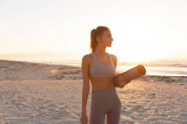 Femme caucasienne tenant tapis de yoga souriant tout en se tenant à la plage. yoga fitness et mode de vie sain concept — Photo de stock