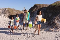 African american parents and two children holding beach accessories walking at the beach. family outdoor leisure time by the sea. — Photo de stock