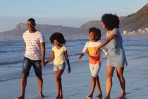Smiling african american parents and two children walking and holding hands at the beach. healthy outdoor leisure time by the sea. - foto de stock