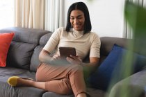 Happy mixed race transgender woman relaxing in living room sitting on couch using tablet. staying at home in isolation during quarantine lockdown. — Stock Photo