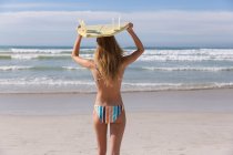 Kaukasische Frau im Bikini mit Surfbrett auf dem Kopf am Strand. gesunde Freizeit im Freien am Meer. — Stockfoto