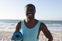 Портрет улыбающегося африканского американца, занимающегося спортом на открытом воздухе, держащего коврик для йоги на пляже. фитнес-тренировки — стоковое фото