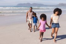 Африканські американські батьки і двоє дітей посміхаються, ходять і тримаються за руки на пляжі. сім'я на відкритому повітрі відпочиває біля моря. — стокове фото