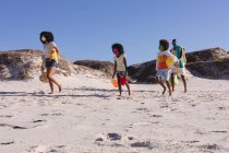 Африканские американские родители и двое детей с пляжными аксессуарами, гуляющими по пляжу. семейный отдых на открытом воздухе у моря во время пандемии коронавируса. — стоковое фото