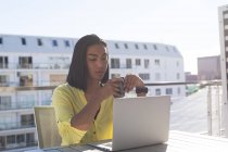 Transgender-Frau mit gemischter Rasse sitzt mit Laptop am Tisch auf sonniger Dachterrasse und trinkt Kaffee. Isolationshaft während der Quarantäne. — Stockfoto