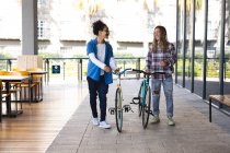 Two happy mixed race male friends wheeling bicycles in the street and talking. green urban lifestyle, out and about in the city. — Stock Photo