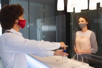 Diverso hombre de negocios con máscara facial desinfectando las manos hablando con la recepcionista en el hotel. viaje de negocios hotel durante coronavirus covid 19 pandemia. - foto de stock