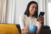 Feliz mujer transexual de raza mixta relajándose en la sala de estar sentada en el sofá usando un teléfono inteligente. permanecer en casa aislado durante el bloqueo de cuarentena. - foto de stock