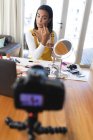 Смешанная расовая трансгендерная женщина делает видеоблог, используя ноутбук и камеру, накладывая макияж. оставаться дома в изоляции во время карантинной изоляции. — стоковое фото