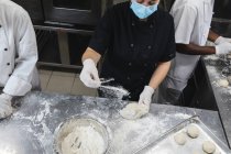 Chefs profesionales de raza mixta que preparan masa con guantes sanitarios y mascarilla facial. trabajando en una ajetreada cocina de restaurante durante coronavirus covid 19 pandemia. - foto de stock