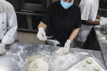 Misturados corrida chefs profissionais preparando massa vestindo luvas sanitárias e máscara facial. trabalhando em uma cozinha restaurante ocupado durante coronavírus covid 19 pandemia. — Fotografia de Stock