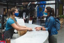 Zwei fröhliche kaukasische Frauen mit Masken reichen eine Tasse Kaffee über den Tresen. Fitness und Freizeit im Fitnessstudio während der Coronavirus-Pandemie 19. — Stockfoto