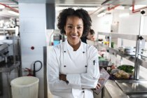 Portrait of smiling mixed race female professional chef with colleagues in background. working in a busy restaurant kitchen. — Stock Photo