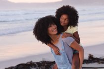 Smiling african american mother carrying her daughter piggyback at the beach. healthy outdoor leisure time by the sea. — Foto stock