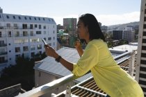 Femme transgenre mixte souriante homme debout sur la terrasse ensoleillée sur le toit tenant du café à l'aide d'un smartphone. rester à la maison dans l'isolement pendant le confinement en quarantaine. — Photo de stock