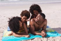 Retrato de padres afroamericanos y dos niños acostados en una toalla en la playa sonriendo. tiempo libre familiar al aire libre junto al mar. - foto de stock