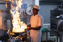 African american male professional chef flambeing dish in wok. working in a busy restaurant kitchen. — Stock Photo