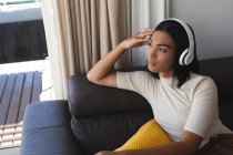 Feliz mestiço transgênero mulher relaxante na sala de estar sentado no sofá com fones de ouvido. ficar em casa em isolamento durante o confinamento de quarentena. — Fotografia de Stock