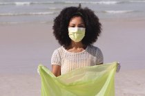 Afrikanisch-amerikanische Frau mit Mundschutz, die Müll am Strand sammelt. Öko-Strandschutz während der Coronavirus-Pandemie. — Stockfoto