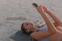 Белая женщина носит наушники, используя смартфон, пока лежит на коврике для йоги на пляже. фитнес-йога и концепция здорового образа жизни — стоковое фото