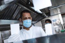 Profi-Koch mit gemischter Rasse schaut sich die Bestellungen mit Hygienehandschuhen und Gesichtsmaske an. Arbeit in einer belebten Restaurantküche während der Coronavirus-Pandemie 19. — Stockfoto