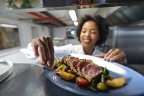 Happy mixed race professional chef finishing dish before serving. working in a busy restaurant kitchen. — Stock Photo