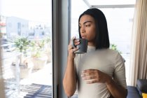 Transgender-Frau mit gemischter Rasse, die aus dem Fenster schaut und eine Tasse Kaffee hält. Isolationshaft während der Quarantäne. — Stockfoto