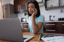 Mujer transgénero de raza mixta que trabaja en casa usando un portátil hablando en un teléfono inteligente. permanecer en casa aislado durante el bloqueo de cuarentena. - foto de stock