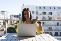 Femme transgenre mixte souriante assise à table sur une terrasse ensoleillée à l'aide d'un ordinateur portable tenant du café. rester à la maison dans l'isolement pendant le confinement en quarantaine. — Photo de stock