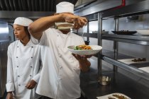 Chef profesional de carrera mixta terminando el plato antes de servir con su colega en segundo plano. trabajando en una cocina ajetreada. - foto de stock