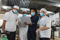 Vielfältige Gruppe professioneller Köche trifft sich mit dem Küchenchef, der Gesichtsmasken trägt. Arbeit in einer belebten Restaurantküche während der Coronavirus-Pandemie 19. — Stockfoto