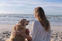 Caucasian woman sitting on sand petting a dog at the beach. healthy outdoor leisure time by the sea. — Stock Photo