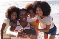 Батьки-афроамериканці та двоє дітей, які роблять селфі зі смартфоном на пляжі, посміхаються. сім'я на відкритому повітрі відпочиває біля моря. — стокове фото